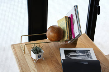 A wooden table features the DETAIL Slope Bookend by DETAIL, a gold magazine rack with books, a wooden sphere, a small succulent in a white planter, and an open photo book showing a black-and-white image.