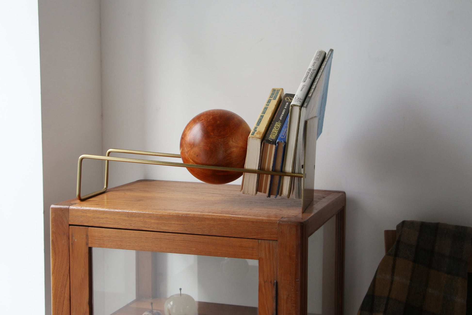 The DETAIL Slope Bookend by DETAIL holds books upright in a modern metal rack on a wooden cabinet against a white wall. Its sculptural, wooden sphere design adds minimalist style to the display.