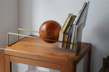 The DETAIL Slope Bookend by DETAIL, with its modern gold metal and large wooden sphere design, supports books on a wooden cabinet against a white wall.