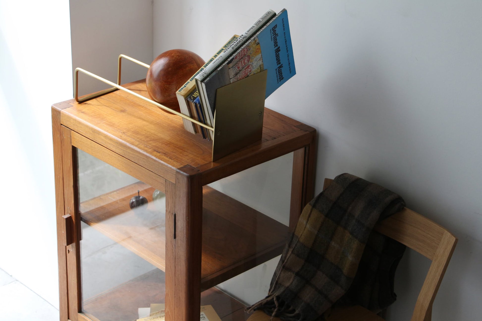 A wooden cabinet with glass doors holds a book on top, propped by the DETAIL Slope Bookend from DETAIL. Next to it, a gold magazine rack contains books and a brown sphere, while a chair nearby has a plaid scarf draped over its backrest.