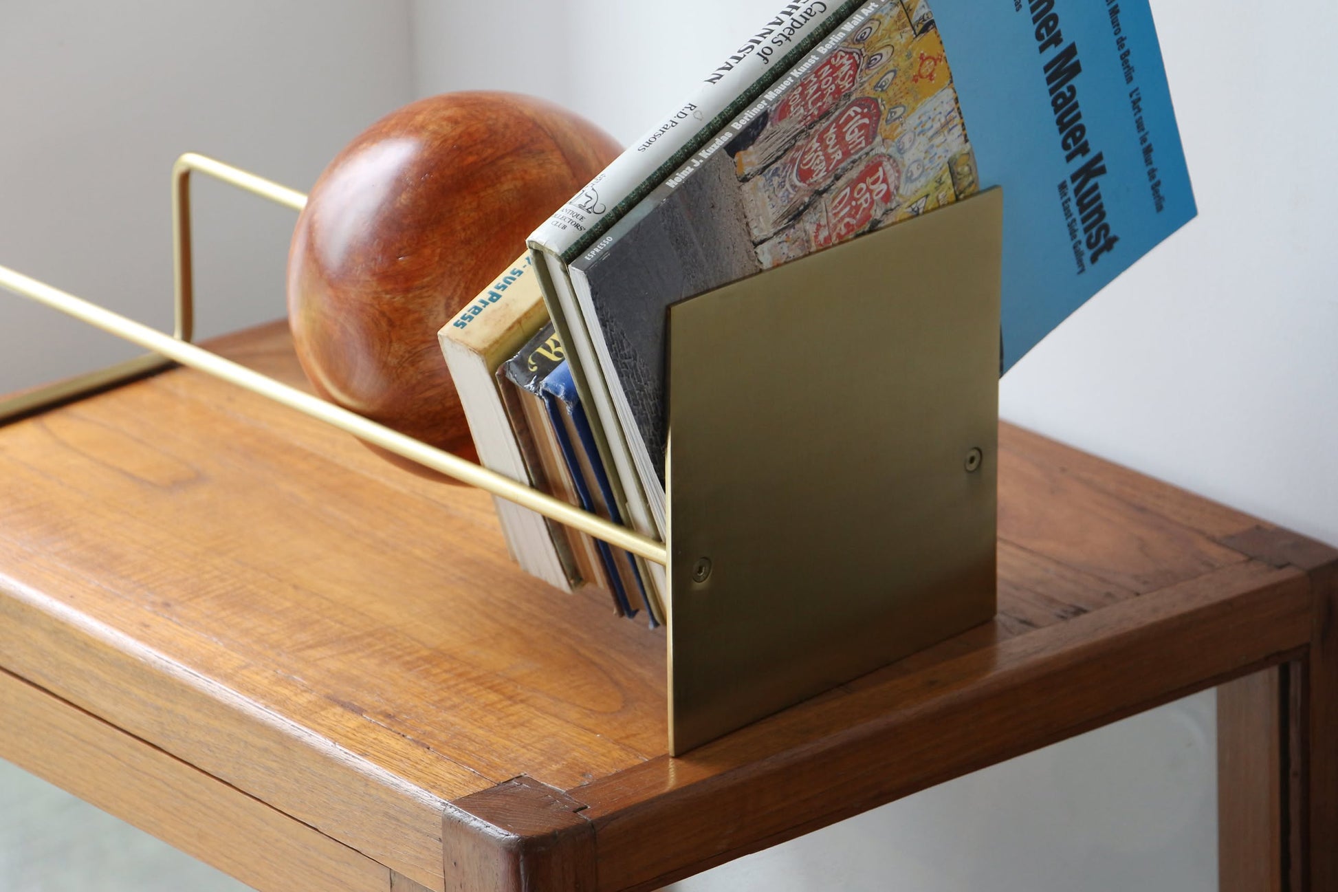 The minimalistic, well-lit scene shows several books upright, anchored by the DETAIL Slope Bookend in modern gold metal from DETAIL, complemented by a large wooden sphere on a wooden side table.