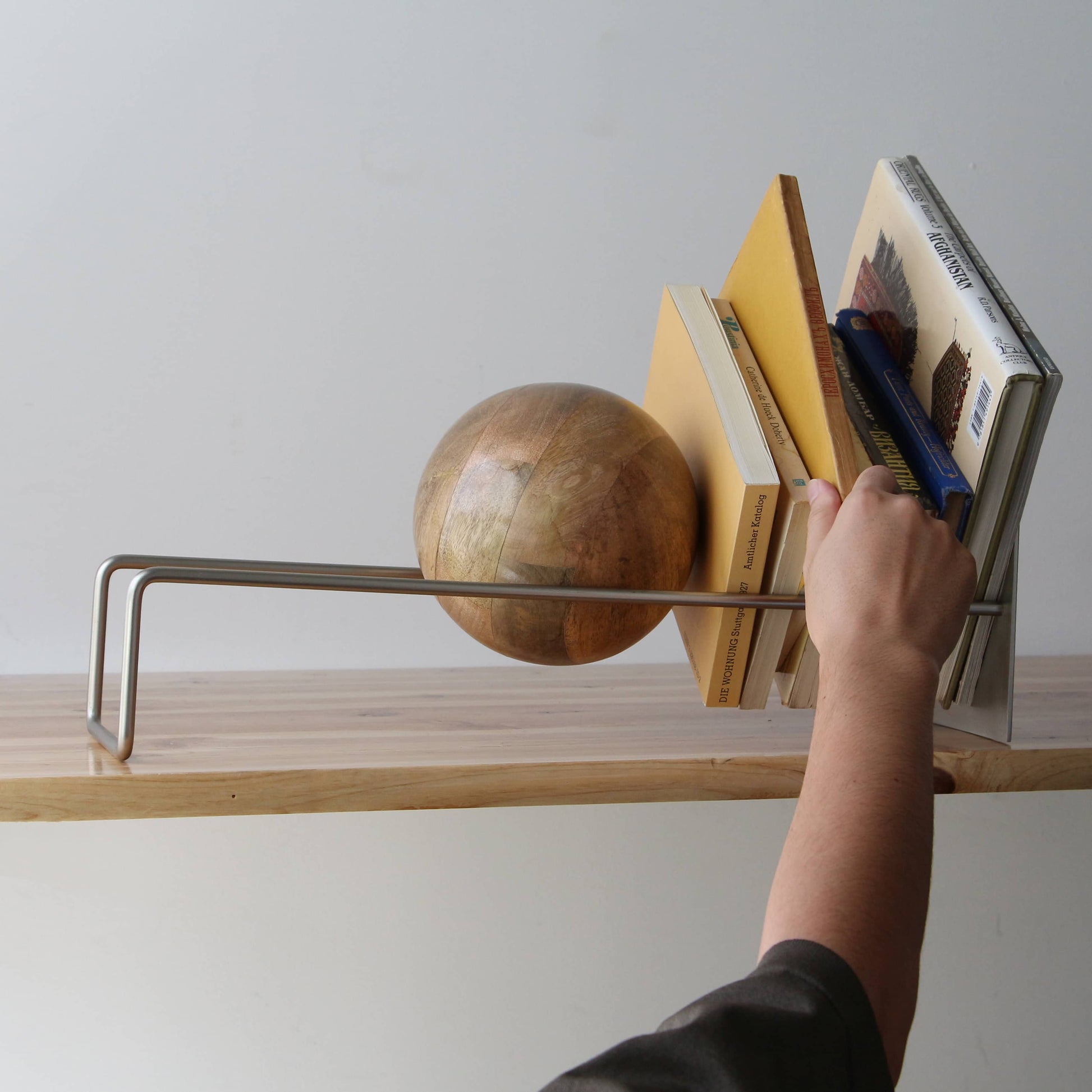A hand adjusts the DETAIL Slope Bookend by DETAIL, a minimalist metal frame with a large wooden sphere that combines form and function to keep books upright on a wooden shelf.