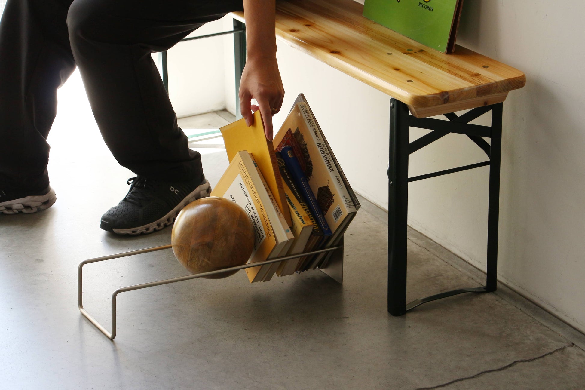 A person seated on a wooden bench reaches for a yellow folder among books and the DETAIL Slope Bookend by DETAIL, with a globe stored in a metal rack on the concrete floor.