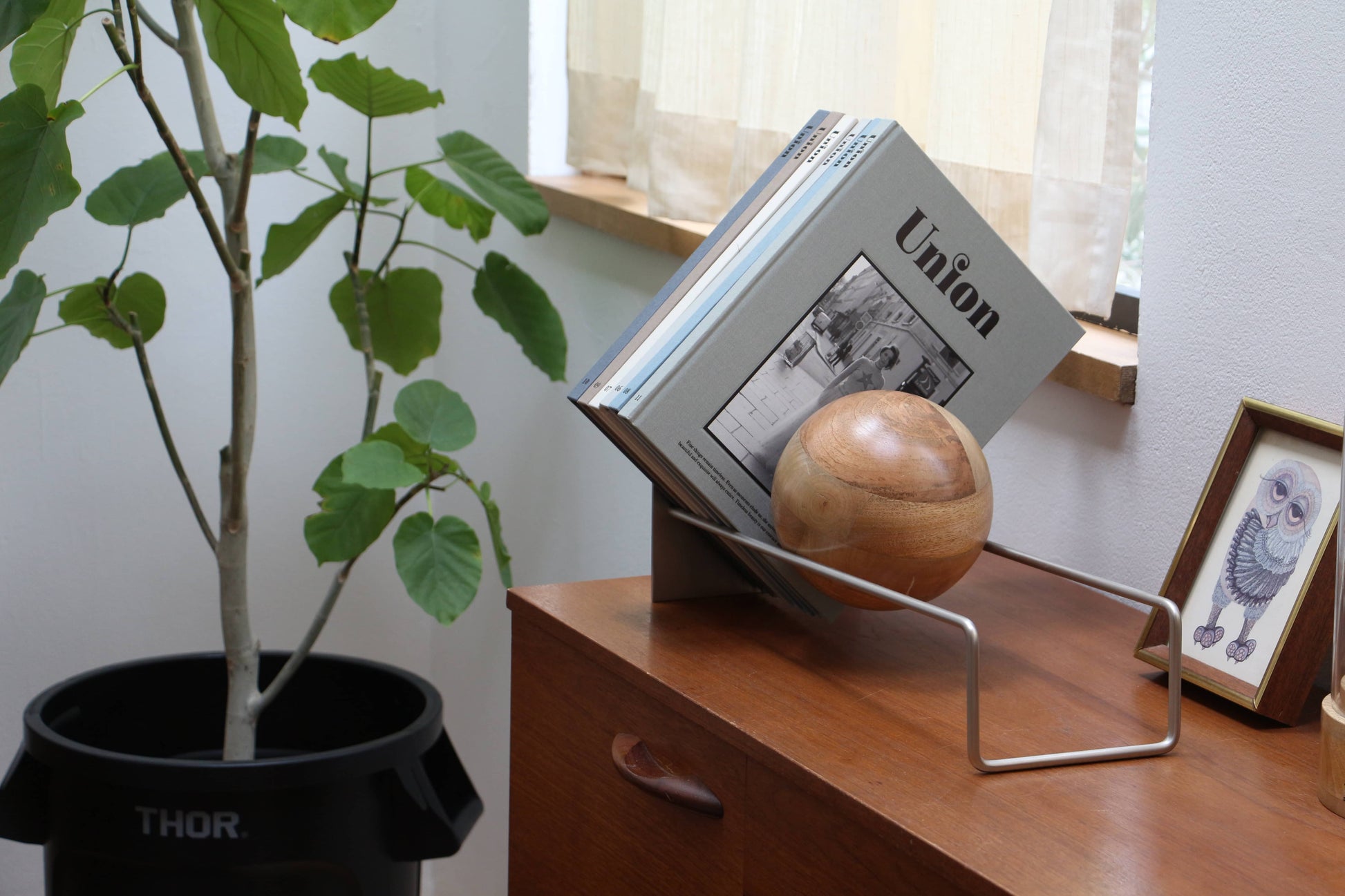 A modern wooden sideboard displays the DETAIL Slope Bookend by DETAIL holding books and a wooden sphere, beside a framed owl illustration and a green potted plant in a black THOR container, with natural light streaming through the window.