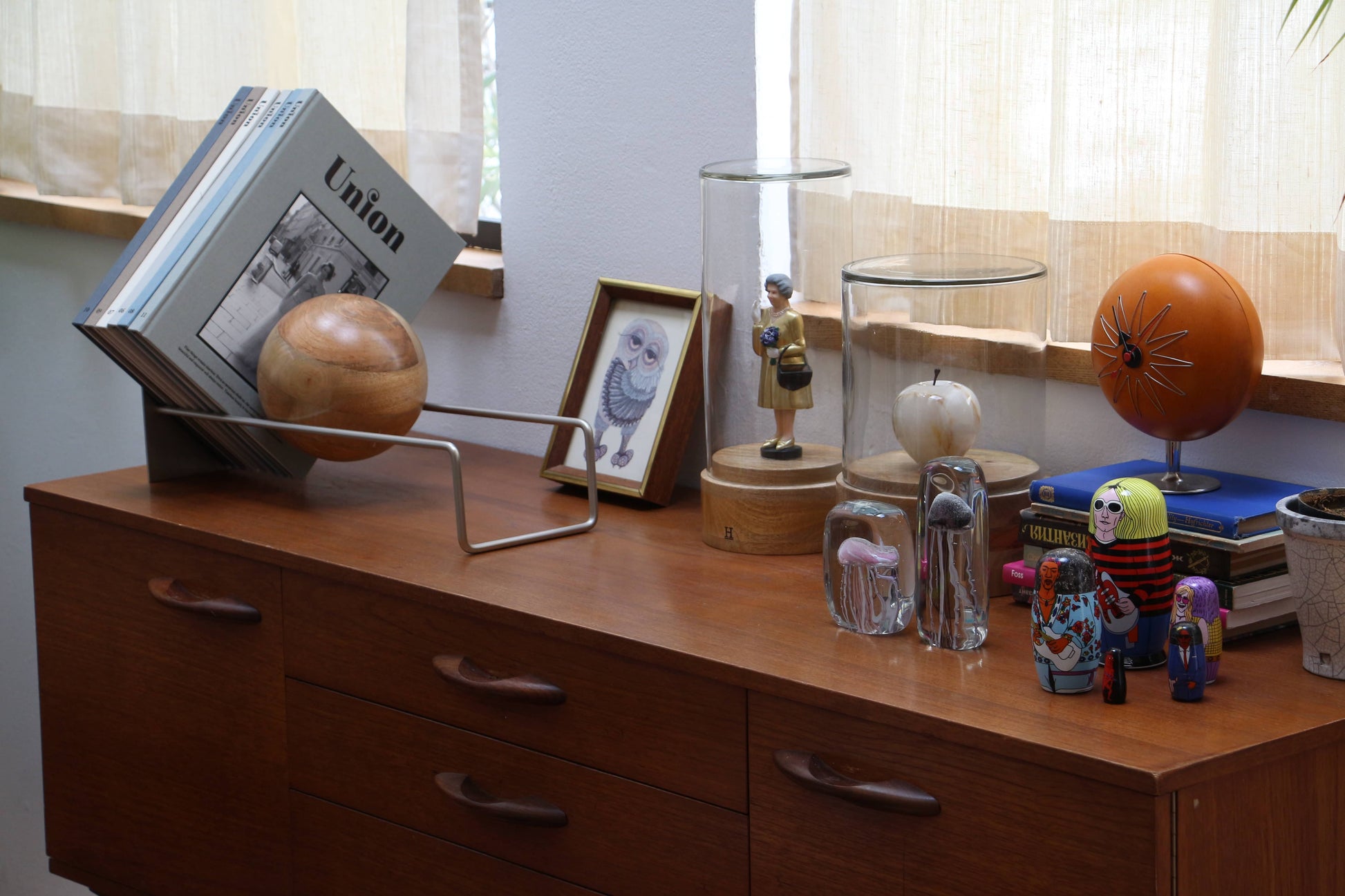 In a sunlit room, stacked magazines, a wooden bowl, framed owl art, glass domes with figurines, a brown globe, and colorful nesting dolls sit atop a wooden sideboard with books neatly arranged by the DETAIL Slope Bookend from DETAIL.