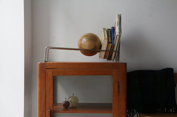 The DETAIL Slope Bookend by DETAIL, a modern metal bookshelf accessory, supports several books and a large wooden sphere on a wooden cabinet that contains three apple-shaped decor pieces; a dark cloth is folded nearby.