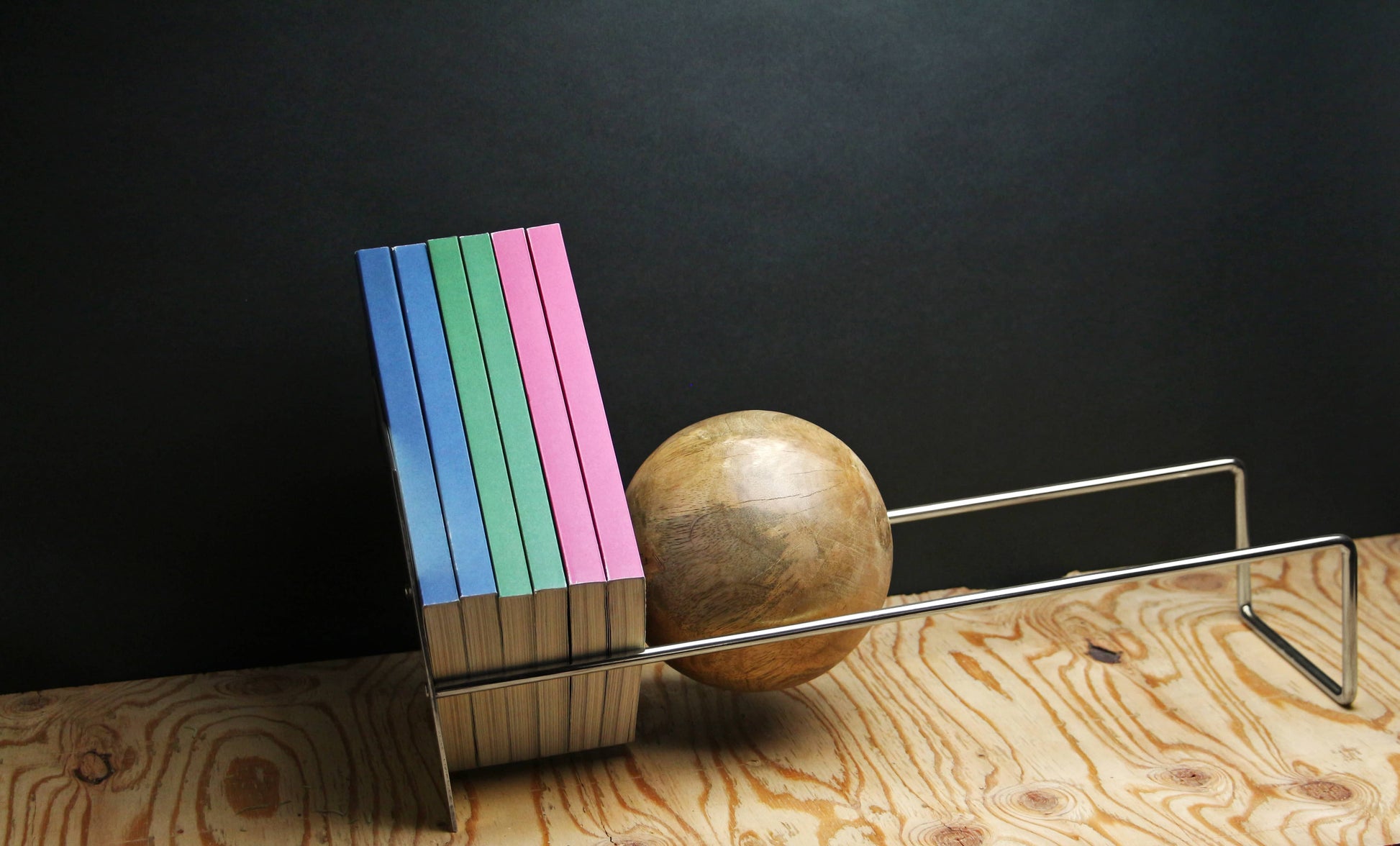 Six pastel-colored books stand upright on a wooden surface, supported by the DETAIL Slope Bookend from DETAIL, alongside metal bookends, set against a dark background.