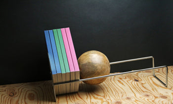 Six pastel-colored books stand upright on a wooden surface, supported by the DETAIL Slope Bookend from DETAIL, alongside metal bookends, set against a dark background.