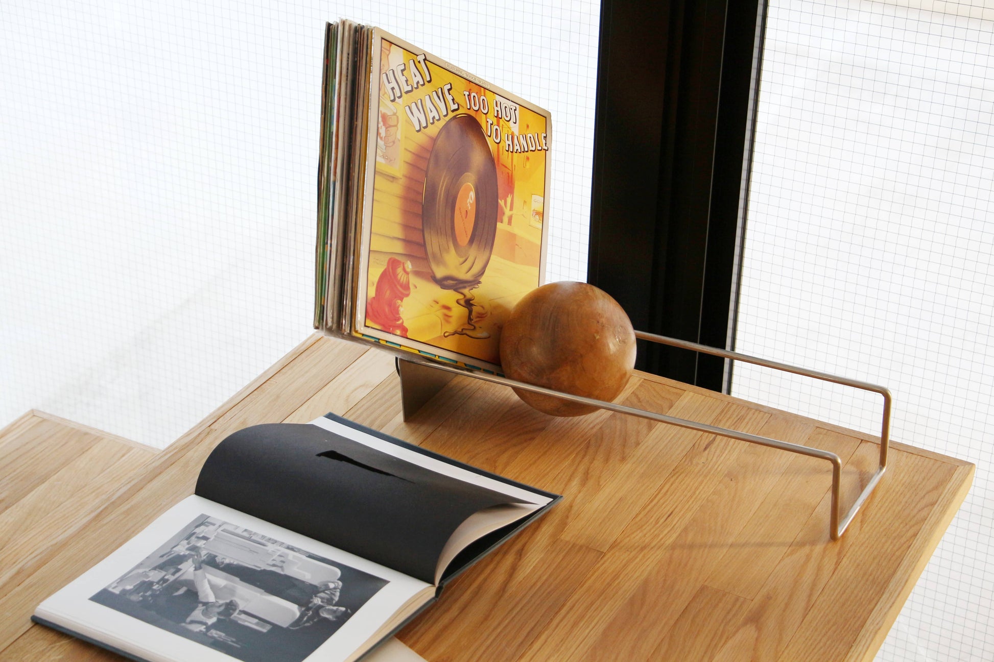 A vinyl record stand holds several albums on a wooden table next to the DETAIL Slope Bookend by DETAIL and an open book displaying black-and-white photos, with sunlight streaming through a window in the background.