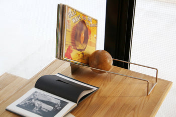 A vinyl record stand holds several albums on a wooden table next to the DETAIL Slope Bookend by DETAIL and an open book displaying black-and-white photos, with sunlight streaming through a window in the background.