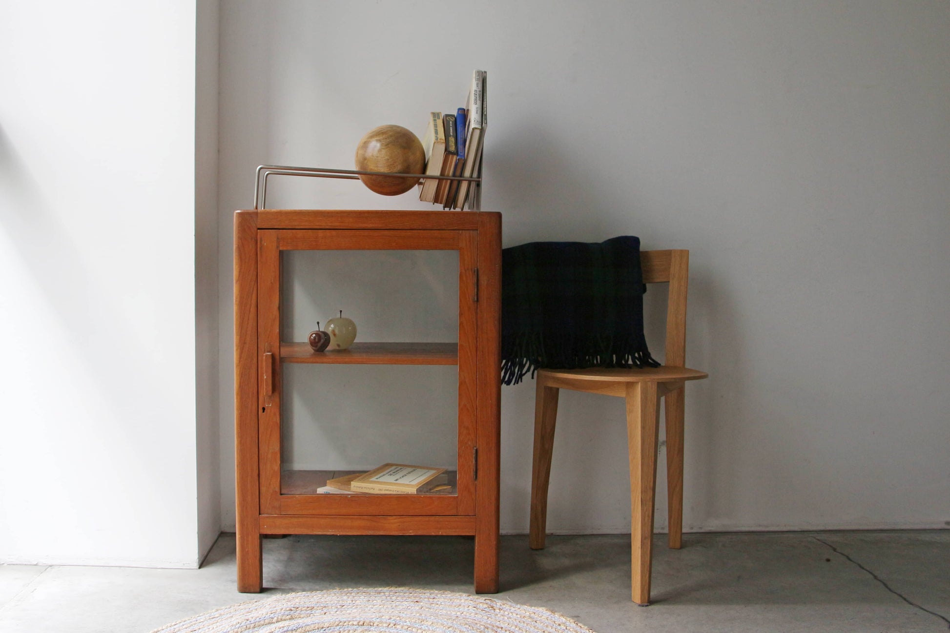 A small wooden cabinet with glass doors stands next to a simple wooden chair draped with a dark plaid blanket. Books, a round wooden sculpture, and the DETAIL Slope Bookend by DETAIL are arranged on and inside the cabinet.