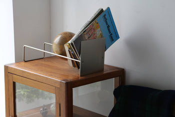 A modern metal magazine holder, books, and magazines sit atop a wooden cabinet next to a chair, featuring the DETAIL Slope Bookend by DETAIL and a decorative wooden sphere behind the holder against a white wall.