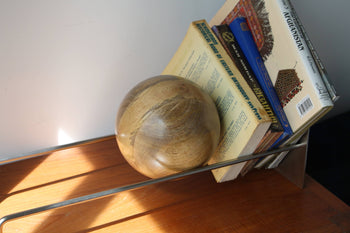 The DETAIL Slope Bookend by DETAIL holds books leaning right on a wooden surface. A polished wooden sphere sits in front, with sunlight casting minimalist shadows, creating a serene scene.