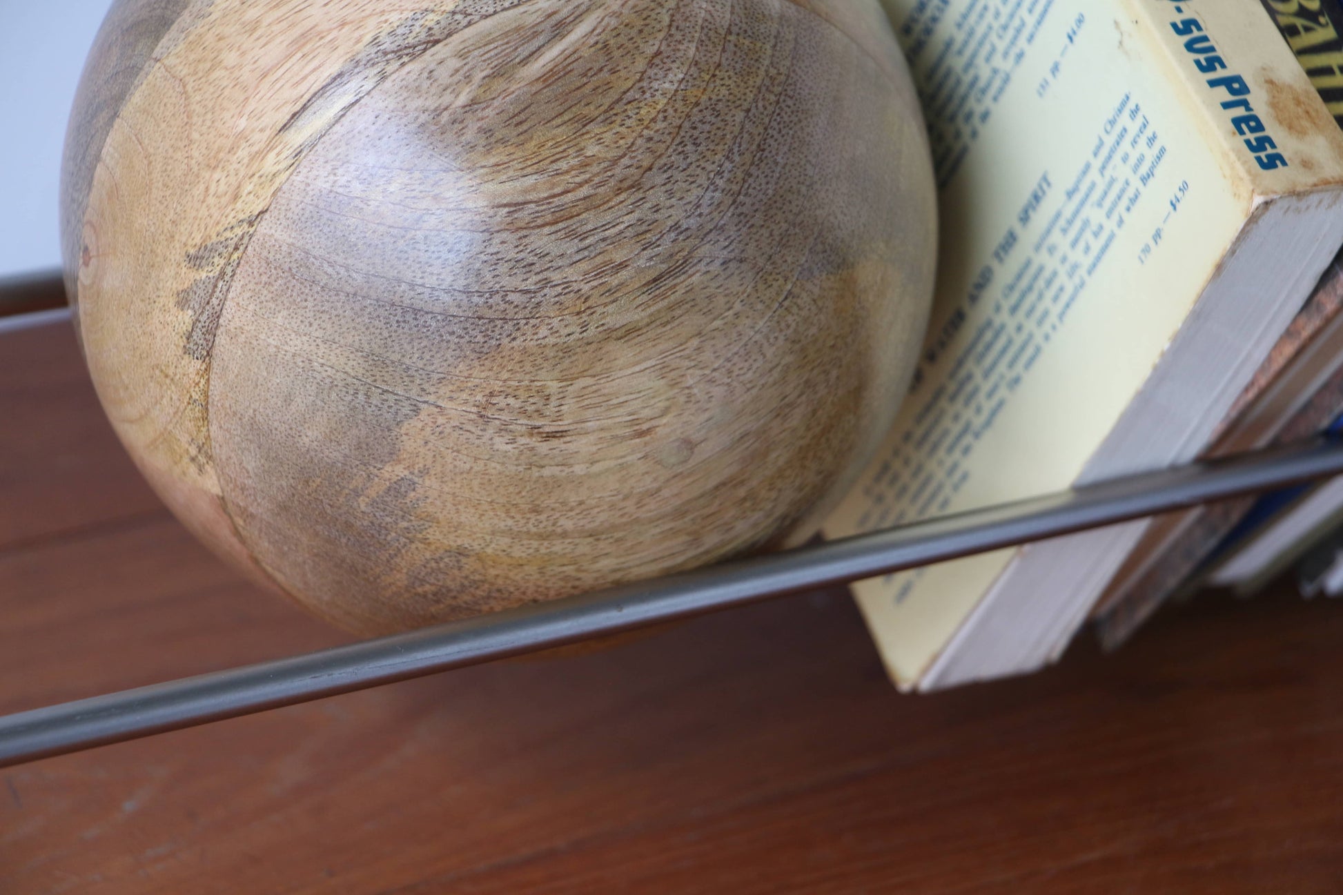 A close-up of the DETAIL Slope Bookend by DETAIL, featuring its sculptural wooden ball design in a metal rack beside books on a wood surface, emphasizing the wood grain and textured book pages.