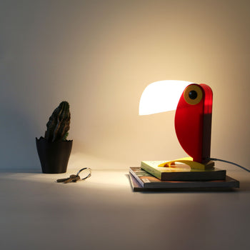 The DETAIL Toucan Lamp by DETAIL, designed in the shape of a red parrot, rests on stacked books and casts light on a cactus in a black pot, with keys on the white tabletop adding vintage flair to the scene.