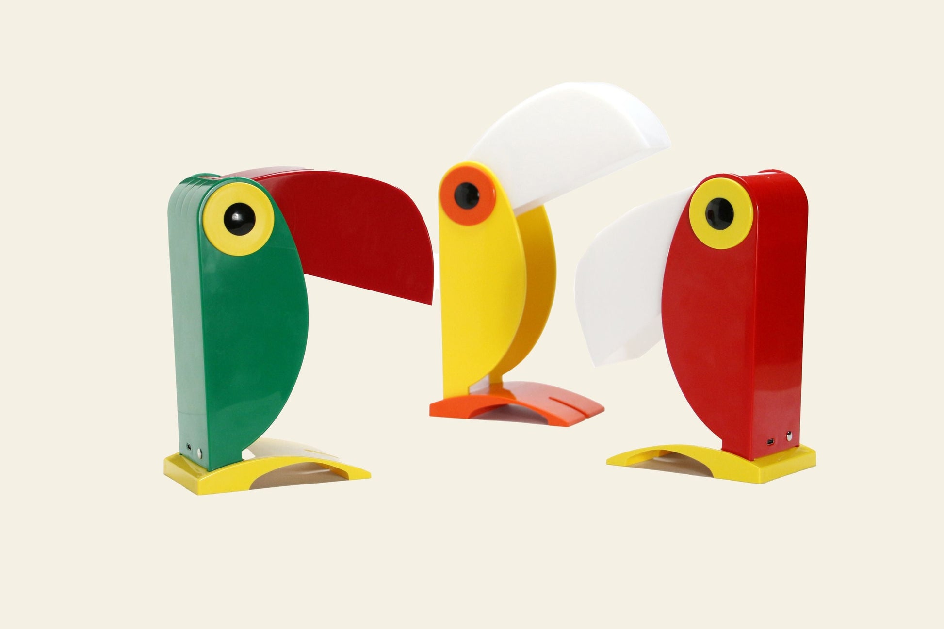 Three DETAIL Toucan Lamps by DETAIL in green, yellow, and red are displayed on a light background. Each lamp features a red or white beak-shaped shade and a geometric body, capturing the vintage charm of this iconic design.