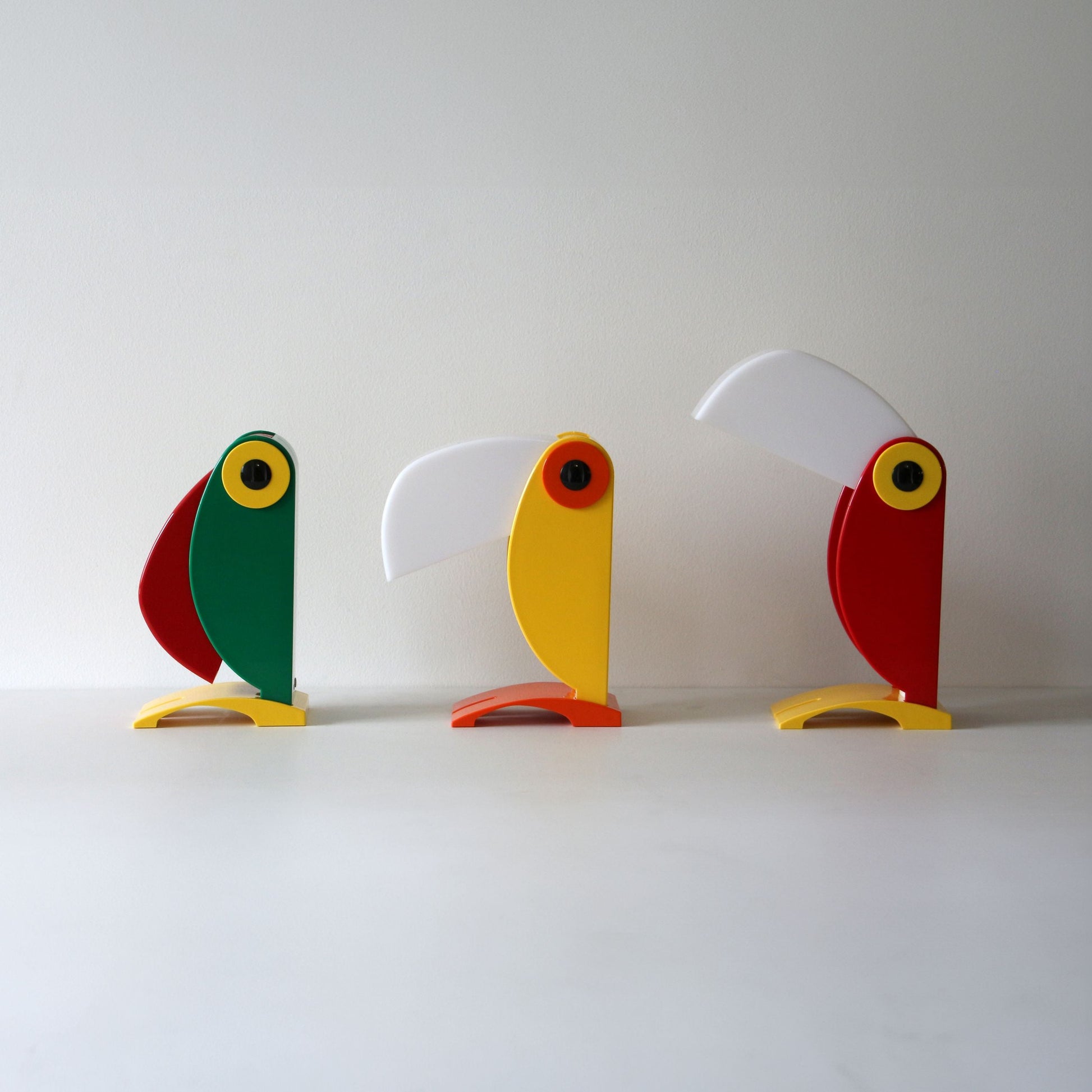 Three minimalist, colorful sculptures sit side by side, their geometric forms and angled beaks recalling the iconic design and vintage charm of the DETAIL Toucan Lamp by DETAIL.
