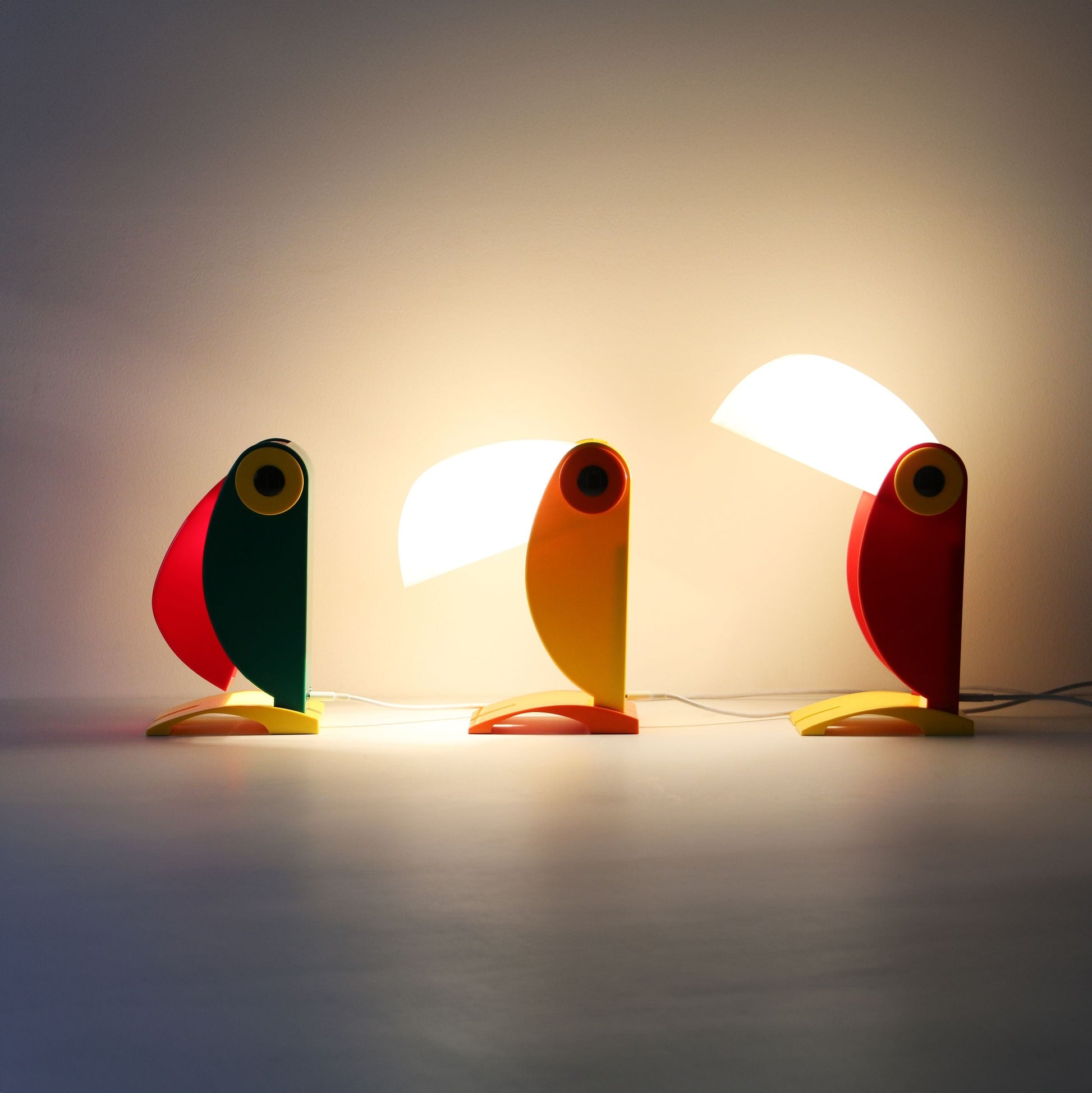 Three DETAIL Toucan Lamps by DETAIL in green, yellow, and red stand in a row with their beaks angled differently. The middle and right lamps are lit, casting a warm glow that highlights their iconic vintage design.