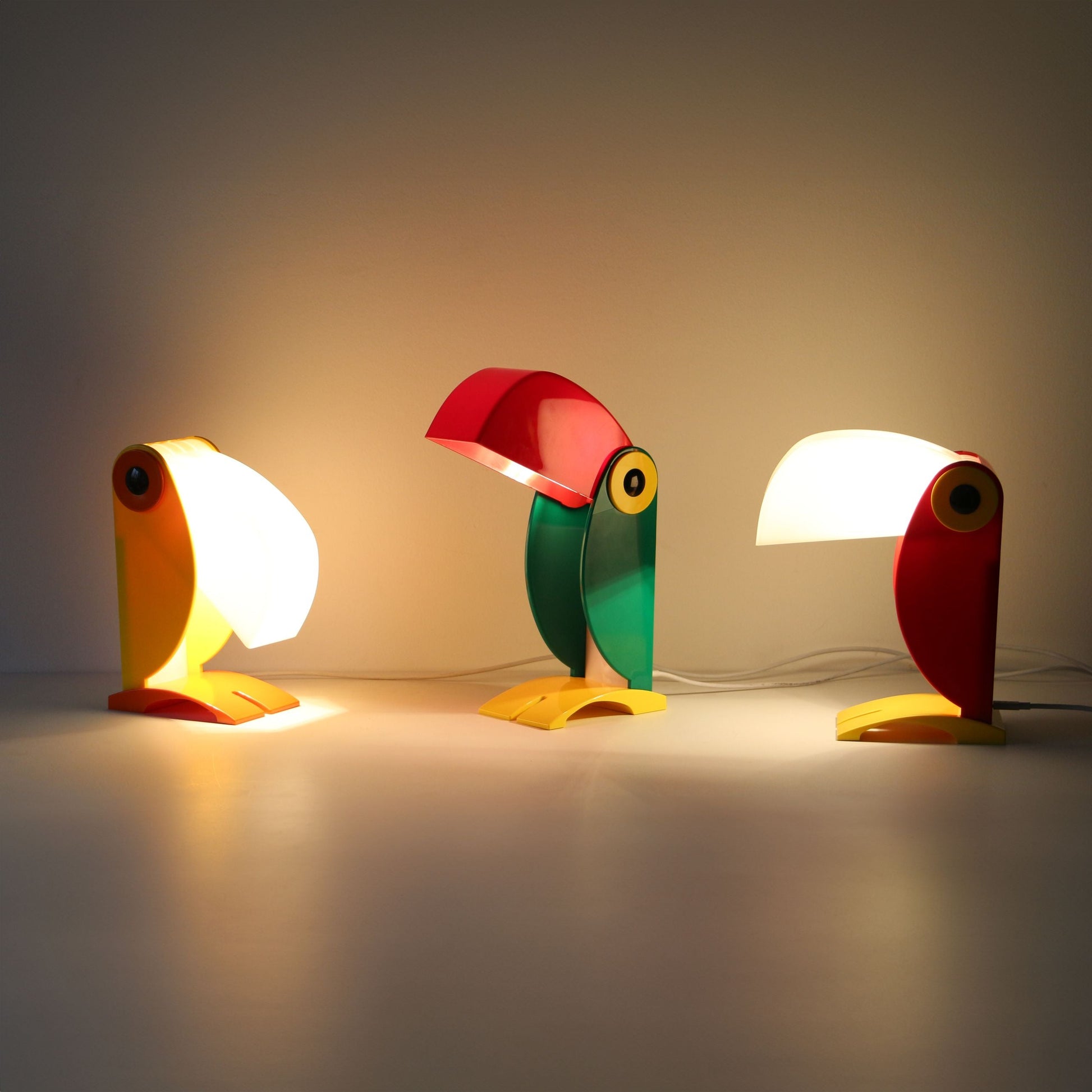 Three DETAIL Toucan Lamps by DETAIL, featuring iconic red, yellow, green, and white designs, are lit on a white surface against a plain light wall—bringing vintage charm to any space.