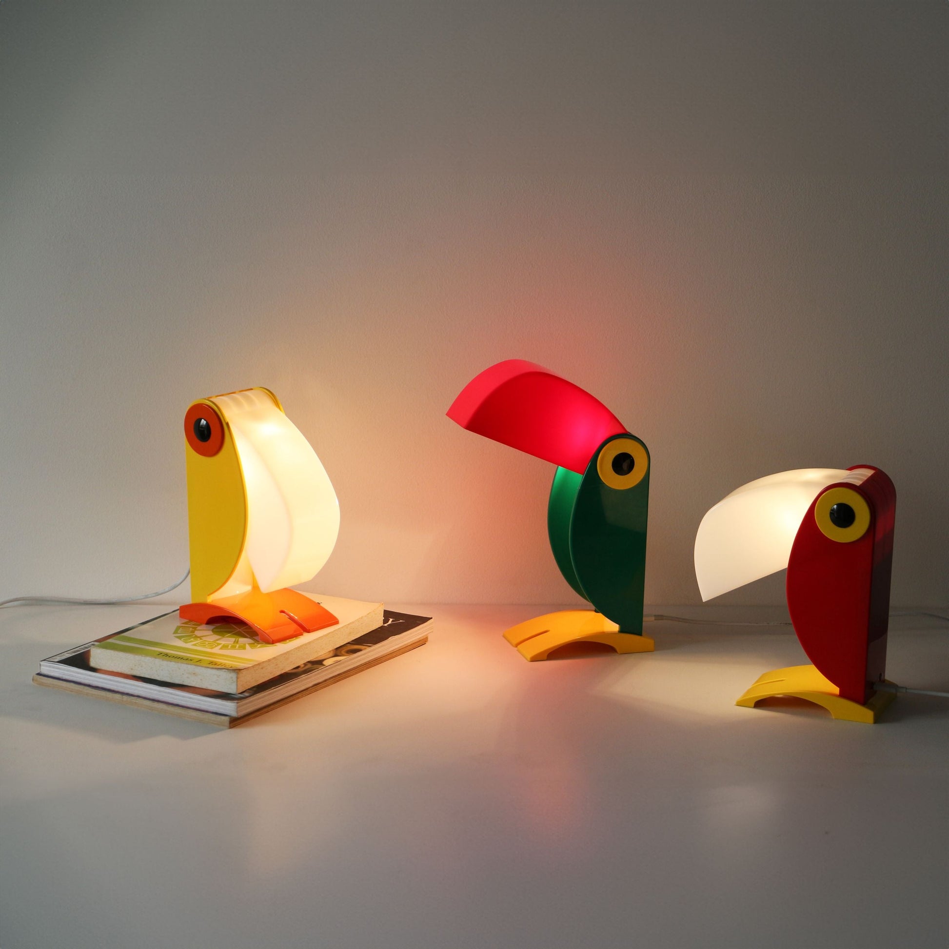Three DETAIL Toucan Lamps by DETAIL, famous for their iconic design and vintage charm, emit a warm glow on a white surface. One lamp rests on stacked magazines, while the other two are placed directly on the table.