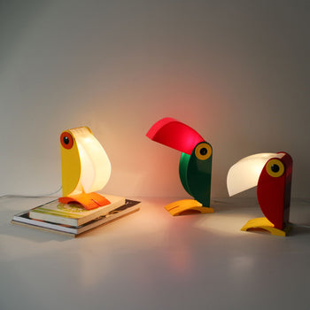 Three DETAIL Toucan Lamps by DETAIL, famous for their iconic design and vintage charm, emit a warm glow on a white surface. One lamp rests on stacked magazines, while the other two are placed directly on the table.