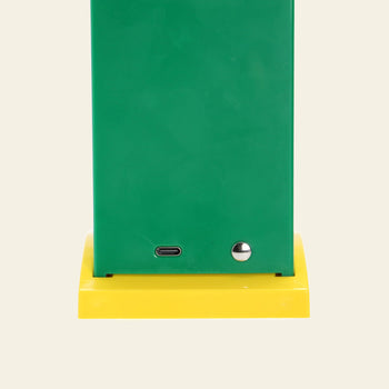 A close-up of the bottom section of the DETAIL Toucan Lamp by DETAIL showcases its green finish, metallic button, and USB-C port, standing on a yellow square base against a plain background to highlight its iconic design.