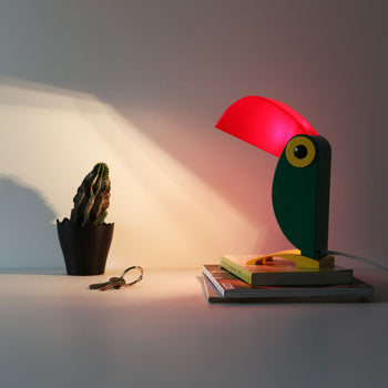 A white desk holds a DETAIL Toucan Lamp by DETAIL emitting red light, a small potted cactus, keys, and a stack of books and notepads against a plain wall.