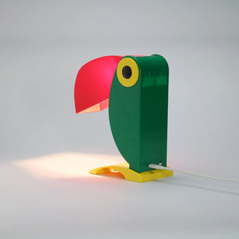 The DETAIL Toucan Lamp by DETAIL features a green body, yellow base and eye, and a red beak lampshade—an iconic vintage design that casts a warm glow onto any white surface.