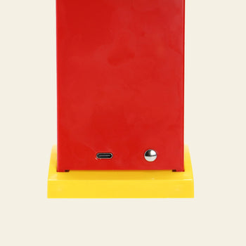 A close-up of the bottom of the red DETAIL Toucan Lamp by DETAIL, featuring a USB-C port and round metal button on a yellow base against a beige background, showcasing its vintage charm.