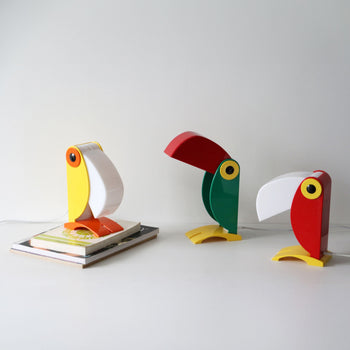 Three colorful DETAIL Toucan Lamps by DETAIL stand on a white surface, each exuding vintage charm—one yellow and white resting on a closed book, one green and red, and one red and white—highlighting their iconic beak-shaped lamp covers.