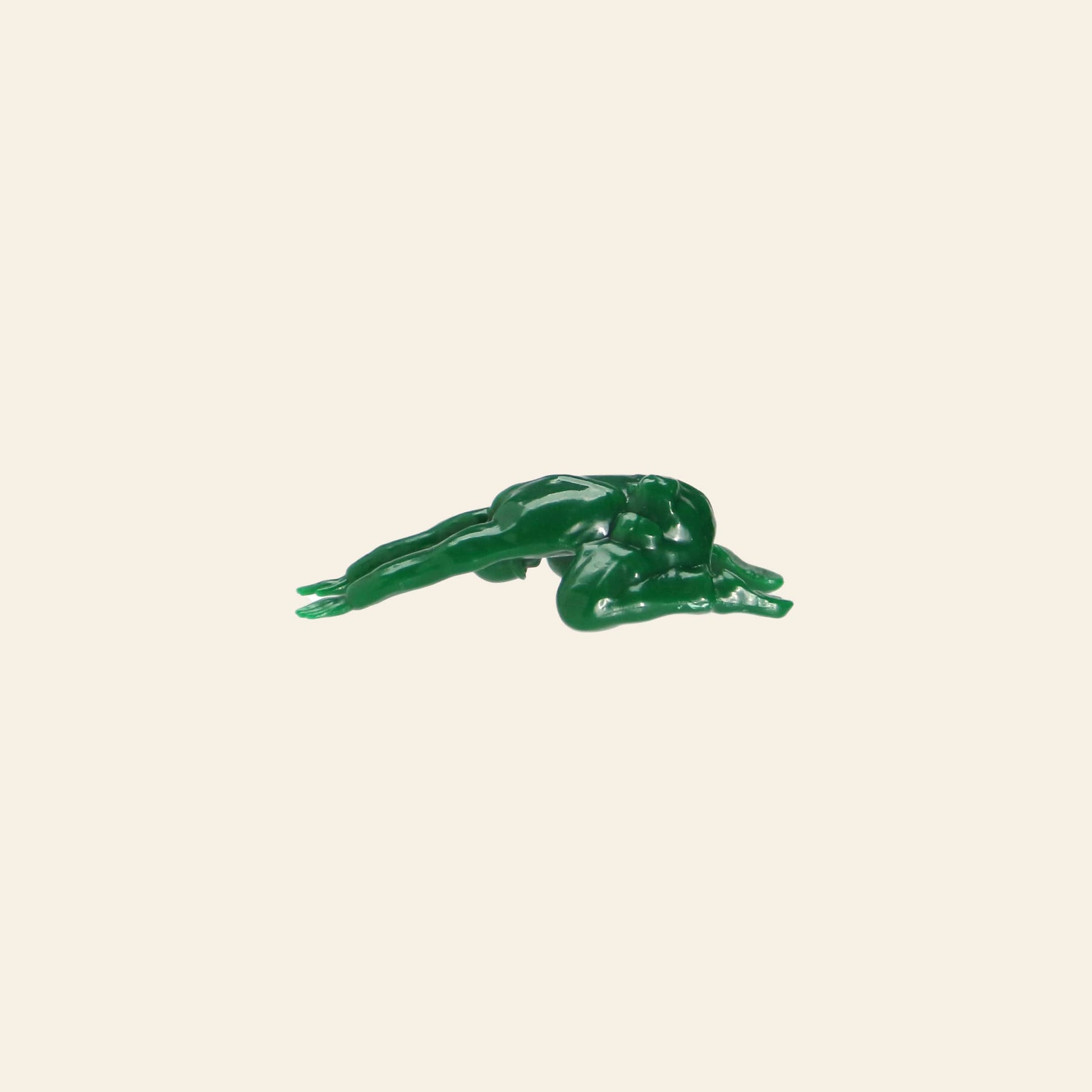 A small green plastic figure from the DETAIL Yoga Joes 9 Figures set by DETAIL lies face down, arms and legs extended like a soldier, against a plain off-white background.