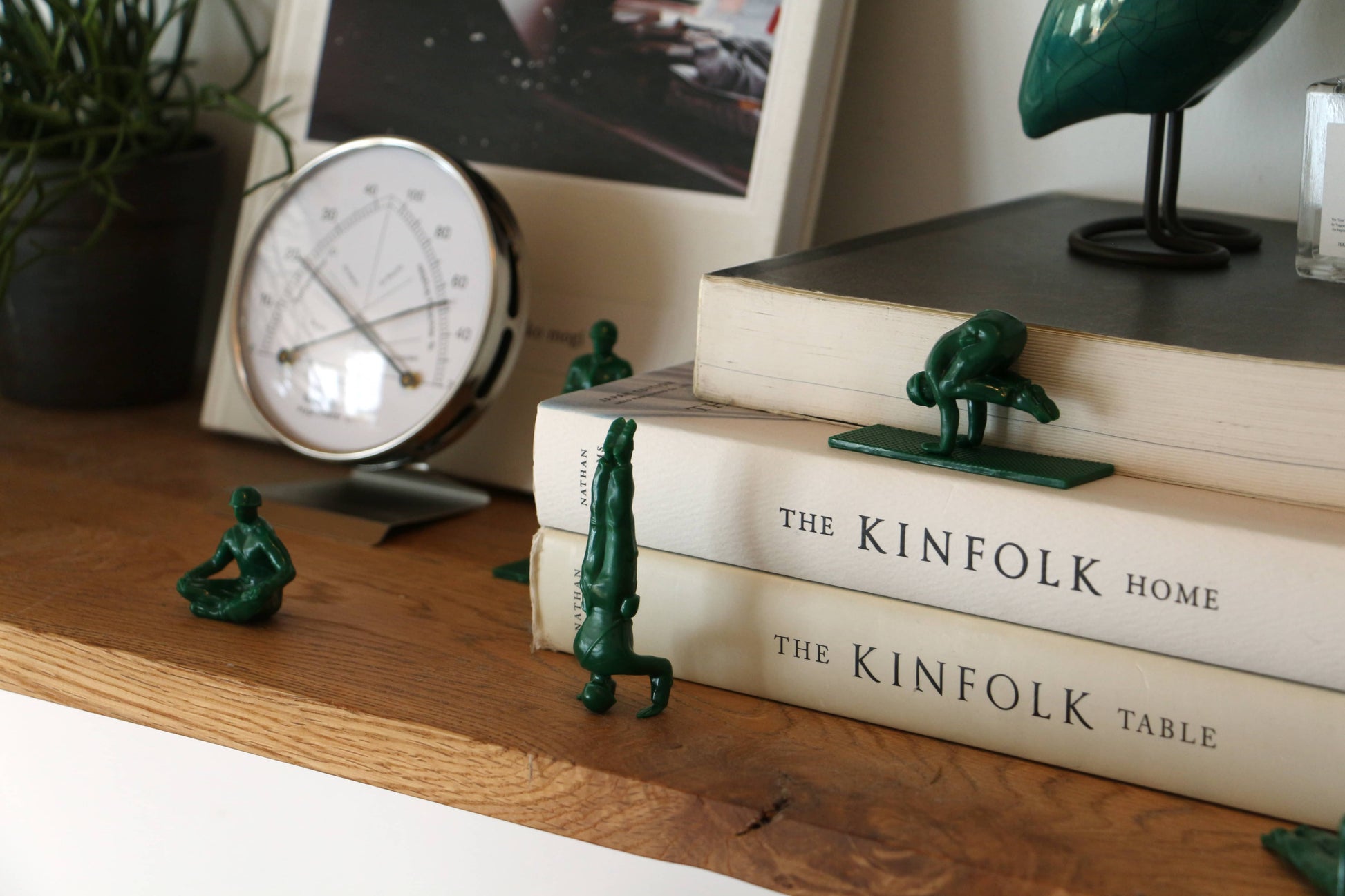 DETAIL Yoga Joes 9 Figures by DETAIL strike playful yoga poses on and around two stacked books, The Kinfolk Home and The Kinfolk Table, displayed on a wooden shelf with a clock, plant, and framed photo in the background.