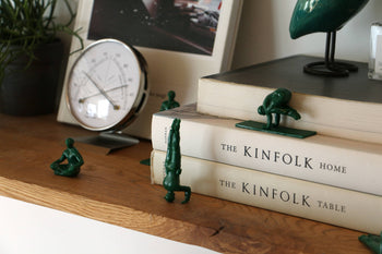 DETAIL Yoga Joes 9 Figures by DETAIL strike playful yoga poses on and around two stacked books, The Kinfolk Home and The Kinfolk Table, displayed on a wooden shelf with a clock, plant, and framed photo in the background.