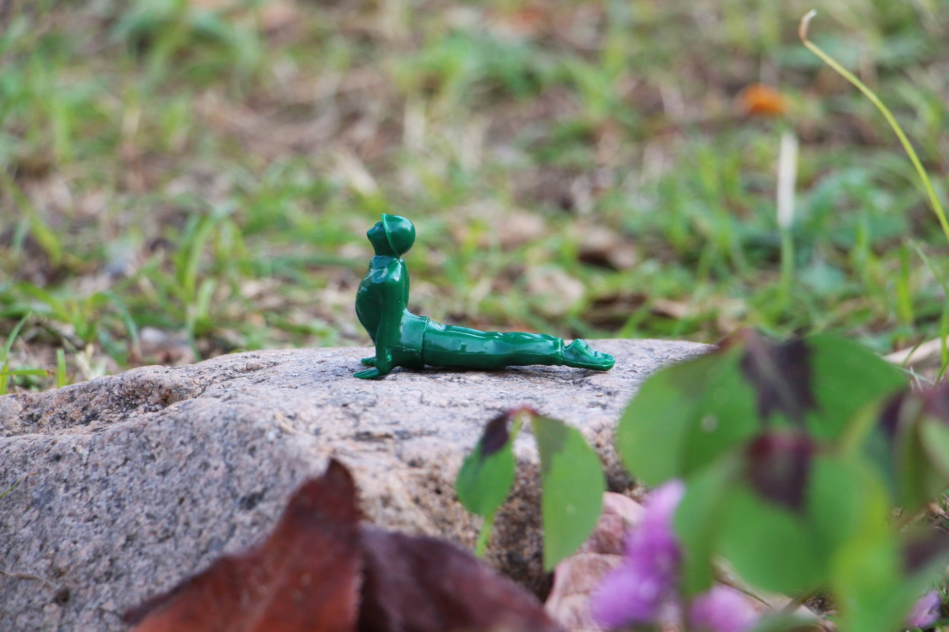A DETAIL Yoga Joes 9 Figures figurine by DETAIL, in a green cobra pose, is set on a rock outdoors with blurred grass and plants in the background.