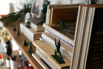 DETAIL Yoga Joes 9 Figures by DETAIL are green plastic toy soldiers posed in yoga positions, arranged on and around stacked books on a wooden shelf, creating a playful and cluttered atmosphere with books and papers.