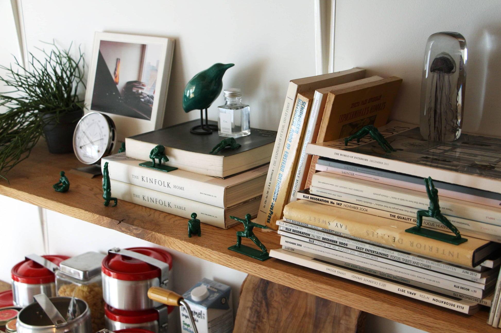 A wooden shelf displays books, DETAIL Yoga Joes 9 Figures by DETAIL in yoga poses, a small clock, framed photo, green bird figurine, silver bulldog bookend, and potted plant. Red kitchen canisters sit below the shelf.