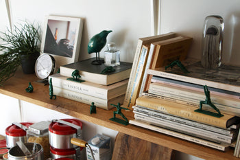 A wooden shelf displays books, DETAIL Yoga Joes 9 Figures by DETAIL in yoga poses, a small clock, framed photo, green bird figurine, silver bulldog bookend, and potted plant. Red kitchen canisters sit below the shelf.