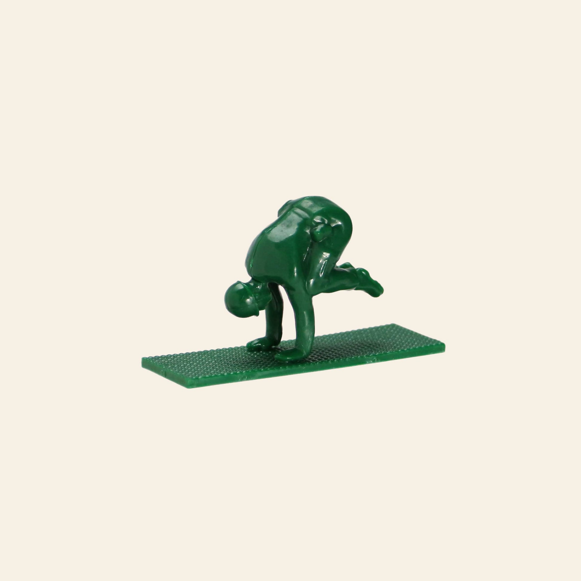 A DETAIL Yoga Joes figure from the 9 Figures set by DETAIL, made of green plastic and mounted on a textured base, balances on its hands with bent legs in a yoga pose against a beige background.