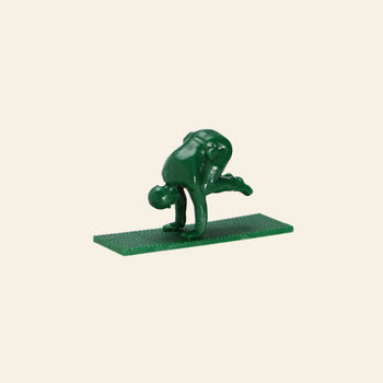 A DETAIL Yoga Joes figure from the 9 Figures set by DETAIL, made of green plastic and mounted on a textured base, balances on its hands with bent legs in a yoga pose against a beige background.