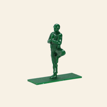 A DETAIL Yoga Joes soldier figure by DETAIL, made of green plastic, stands in the yoga tree pose with arms folded and one foot on the opposite leg atop a rectangular base, shown against a plain beige background. Part of the 9 Figures set.