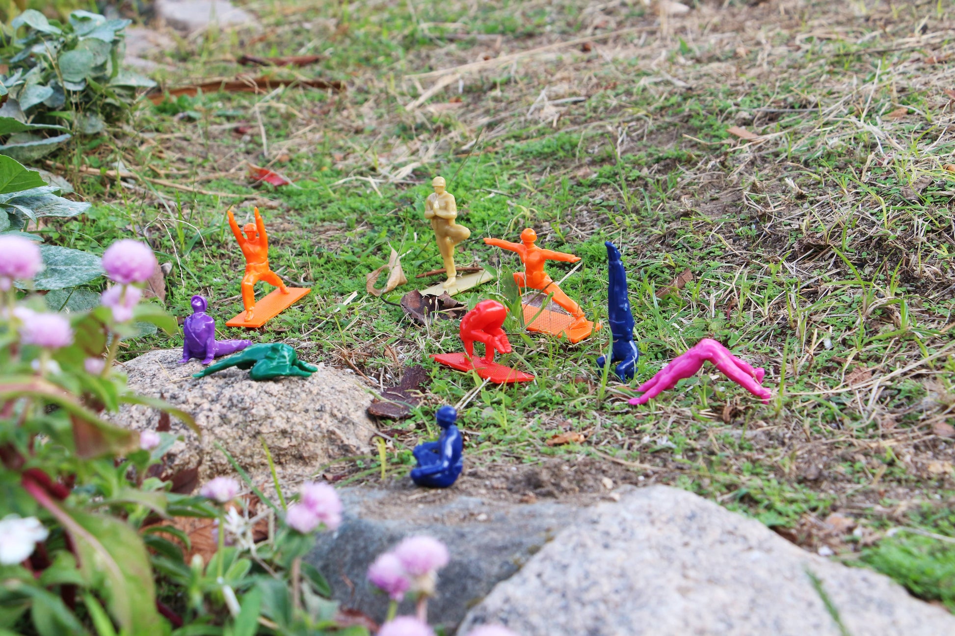 DETAIL Yoga Joes 9 Figures by DETAIL are arranged in yoga poses on grass and rocks in a garden, with pink flowers and green plants in the foreground.