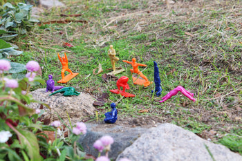 DETAIL Yoga Joes 9 Figures by DETAIL are arranged in yoga poses on grass and rocks in a garden, with pink flowers and green plants in the foreground.