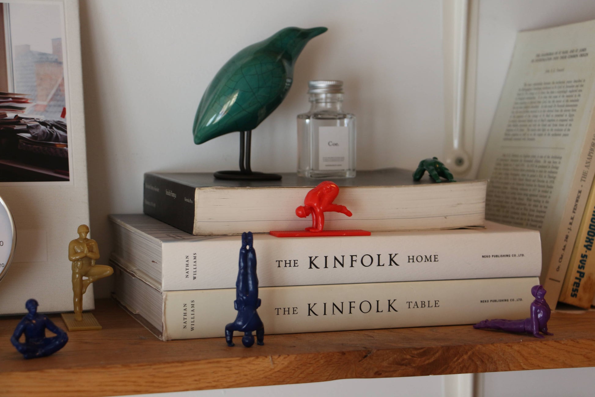 A bookshelf displays DETAIL Yoga Joes 9 Figures in playful yoga poses, books like The Kinfolk Home and The Kinfolk Table, a green bird sculpture, a perfume bottle, and an open book propped in the background.