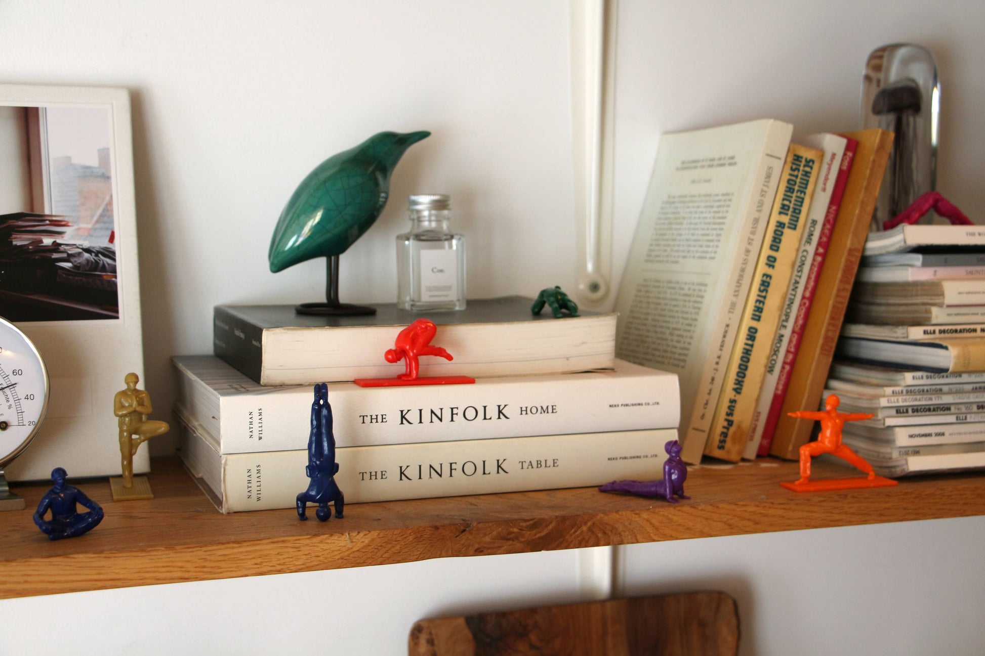 A wooden shelf with books, stacked magazines, colorful DETAIL Yoga Joes 9 Figures by DETAIL, a green bird sculpture, a small bottle, and a framed photo leaning against the wall creates a cozy and eclectic scene.