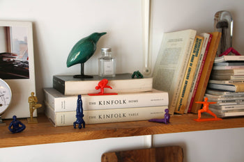 A wooden shelf with books, stacked magazines, colorful DETAIL Yoga Joes 9 Figures by DETAIL, a green bird sculpture, a small bottle, and a framed photo leaning against the wall creates a cozy and eclectic scene.