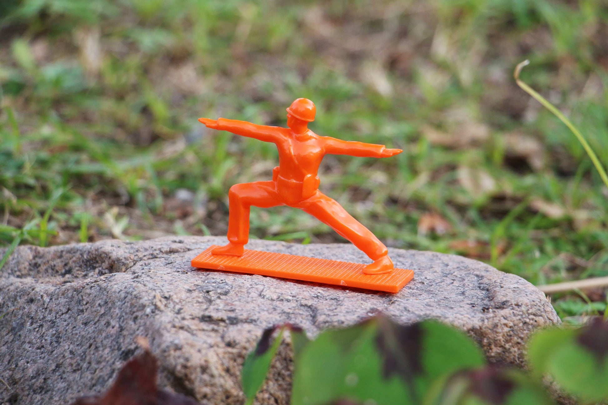 A DETAIL Yoga Joes figurine from the DETAIL Yoga Joes 9 Figures set, in orange and a warrior pose, stands on a textured rock outdoors with green grass and plants in the background.