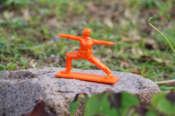 A DETAIL Yoga Joes figurine from the DETAIL Yoga Joes 9 Figures set, in orange and a warrior pose, stands on a textured rock outdoors with green grass and plants in the background.