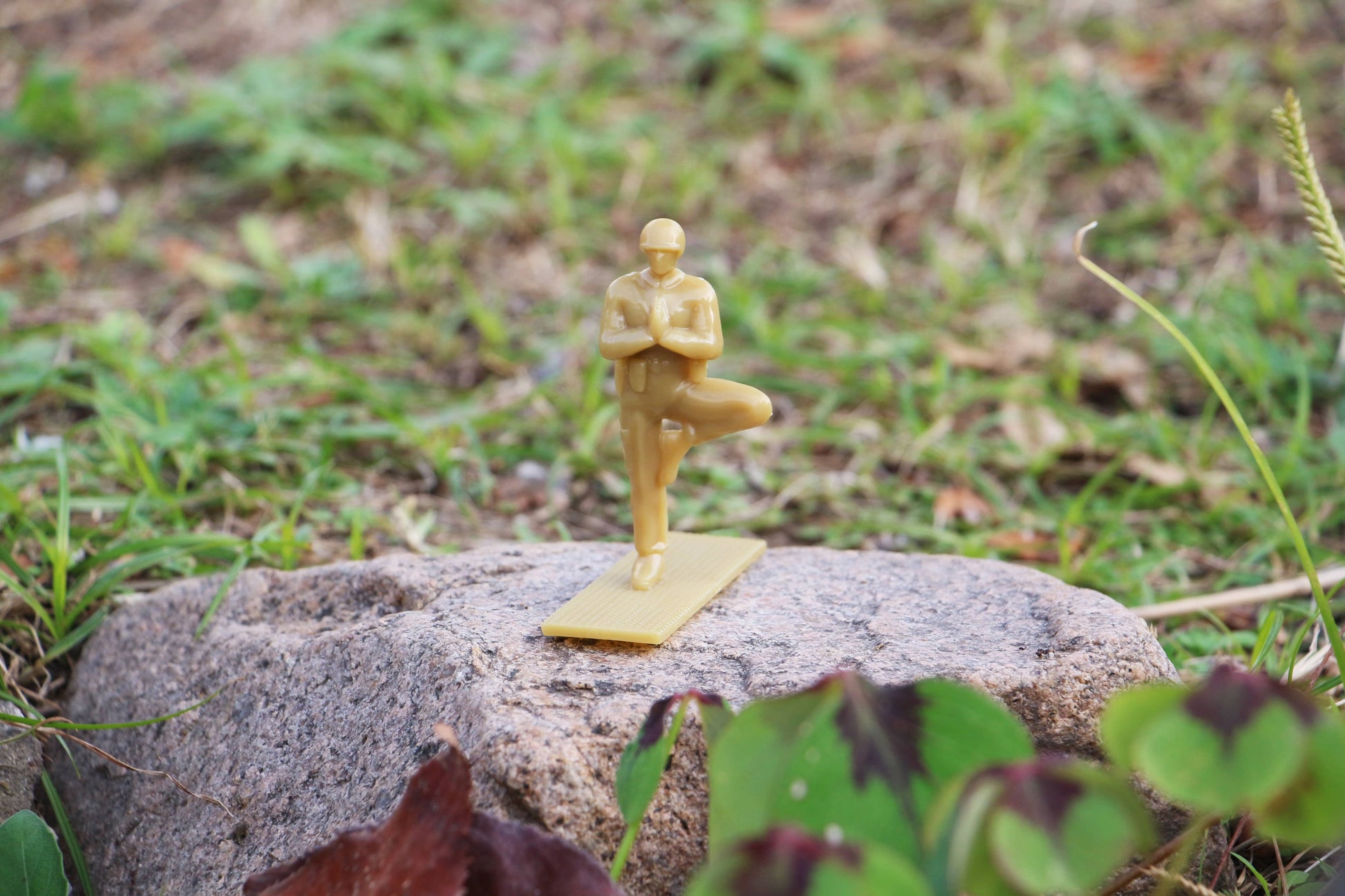 A DETAIL Yoga Joes figure from the DETAIL Yoga Joes 9 Figures set stands in tree pose on a rectangular base atop a rock, with grass and plants in the background.