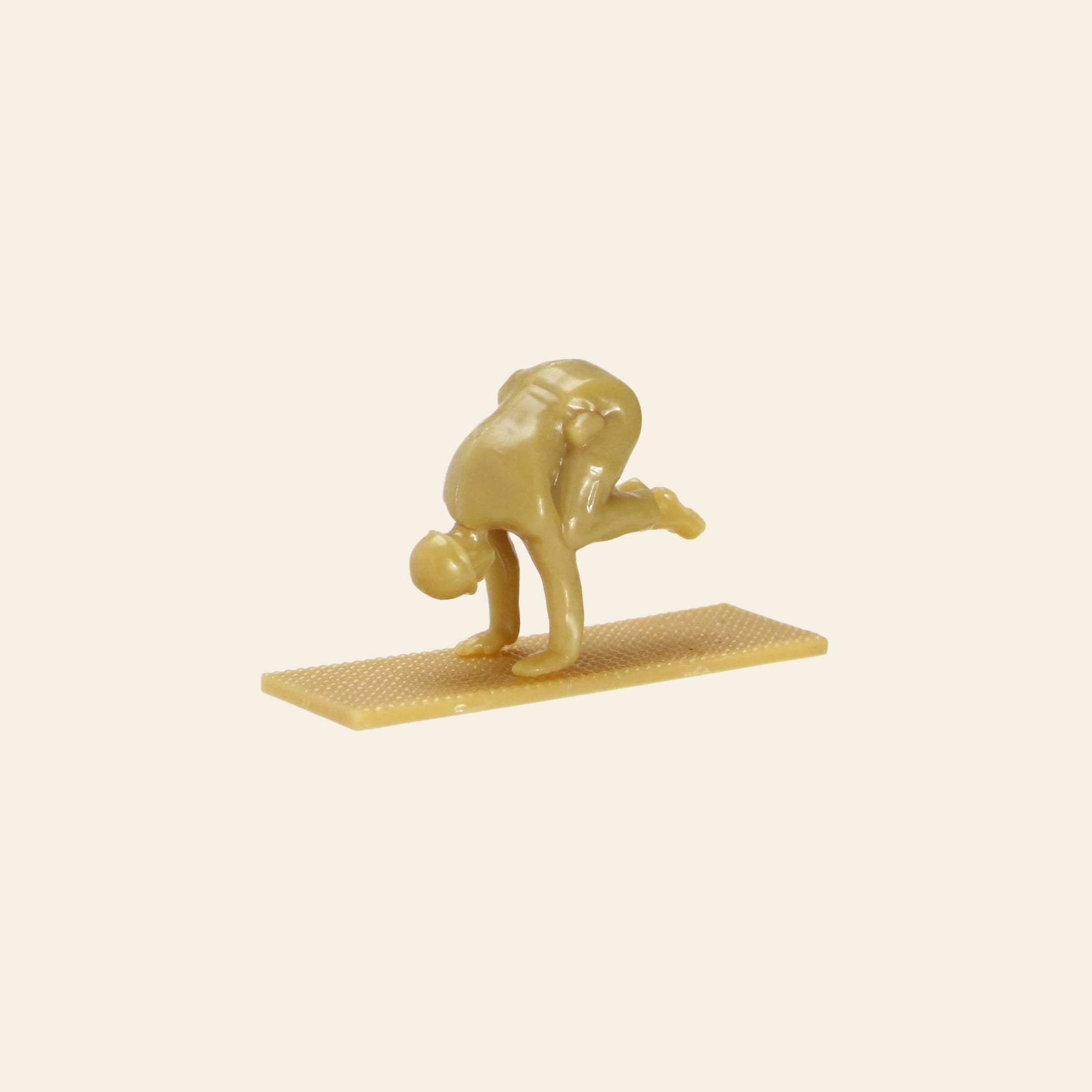 A small, tan DETAIL Yoga Joes 9 Figures figurine from DETAIL is balanced on a rectangular base, bending forward with both hands on the ground and one leg bent upward in a yoga pose.