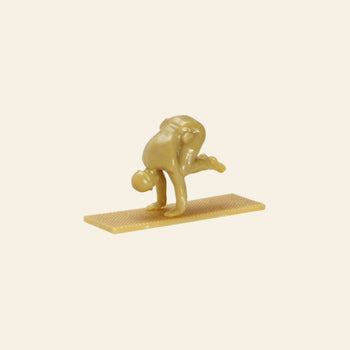 A small, tan DETAIL Yoga Joes 9 Figures figurine from DETAIL is balanced on a rectangular base, bending forward with both hands on the ground and one leg bent upward in a yoga pose.
